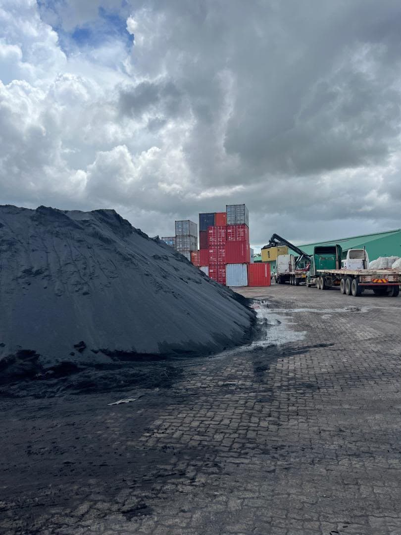 A stockpile of chrome ore at a container terminal, with stacked containers and flatbed trailers behind it